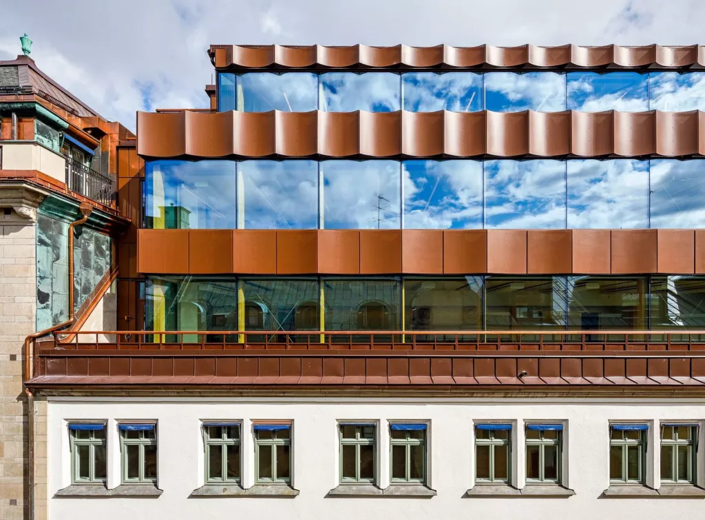 En modern byggnad med en fasad av glas och koppar, där glaset reflekterar en blå himmel med moln.