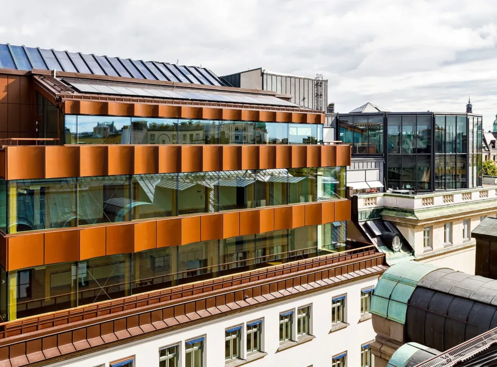 En modern byggnad med orangea fasadpaneler och stora glasfönster, omgiven av äldre arkitektur.