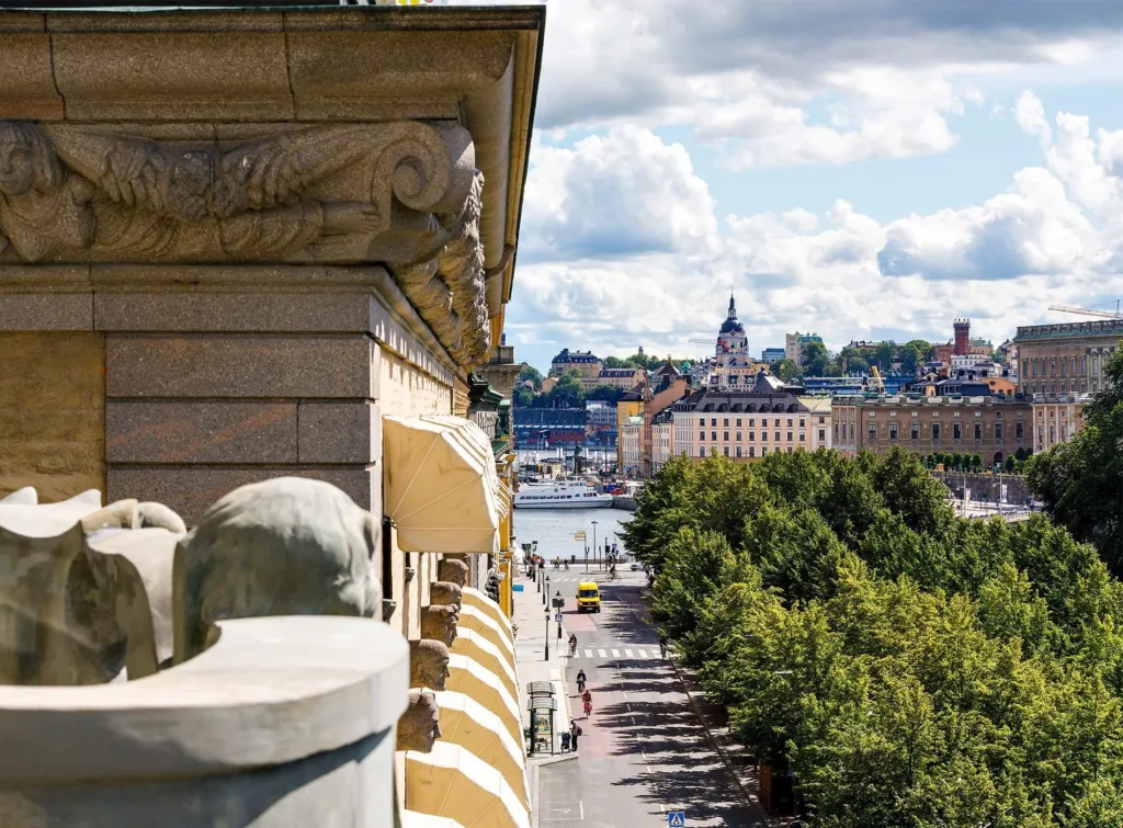 En utsikt över en stadsgata med gröna träd, klassiska byggnader och en kyrka i bakgrunden, sett från en ornamenterad fasad.