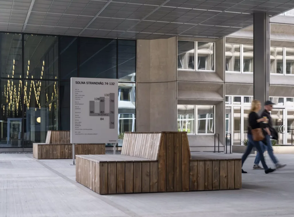 4. Träbänkar och en informationsskylt på en öppen betongyta framför en glasfasad.