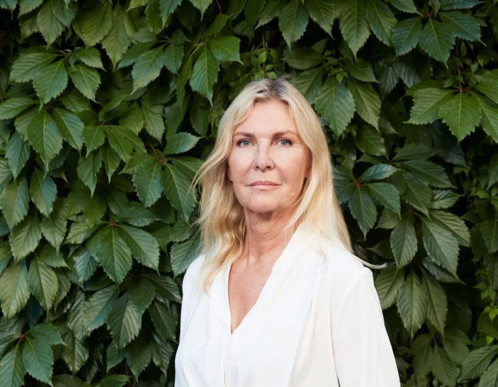 En kvinna med blont hår i vit blus står framför en grön växtvägg.