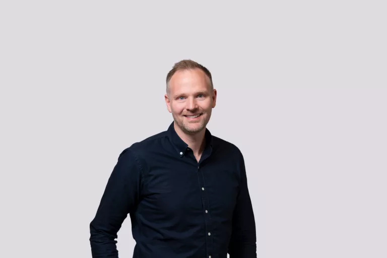 En man med kort hår, klädd i en mörk skjorta, ler mot kameran med en ljus bakgrund.