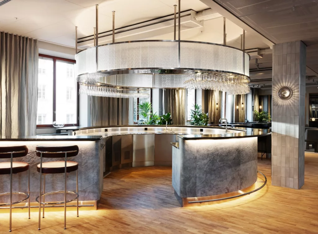 En modern bar med en rund bardisk i metall och glas, omgiven av speglar och trädetaljer.