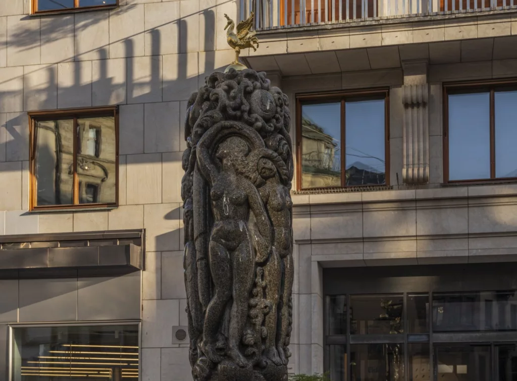En skulptur med detaljerade figurer och en gyllene tupp på toppen, placerad framför en byggnad.