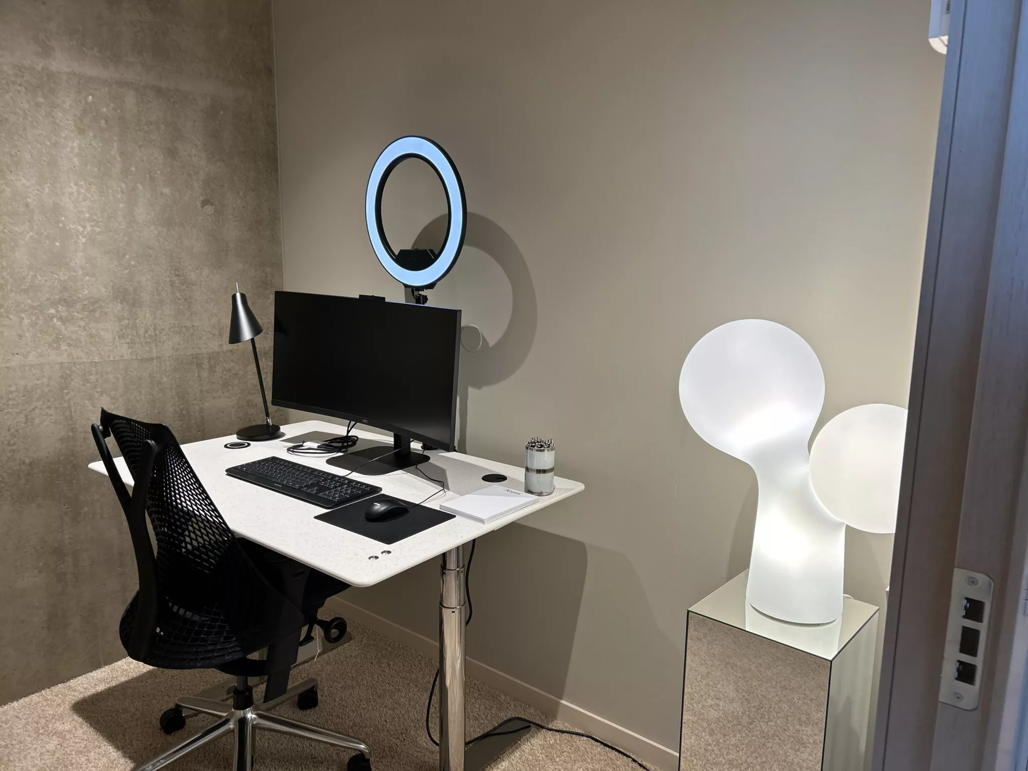 En arbetsplats med ett skrivbord, dator, stol och en cirkelformad lampa på väggen; bredvid står en modern vit lampa på en piedestal.