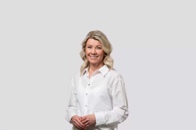 En kvinna med blont hår i en vit skjorta står och ler mot kameran.