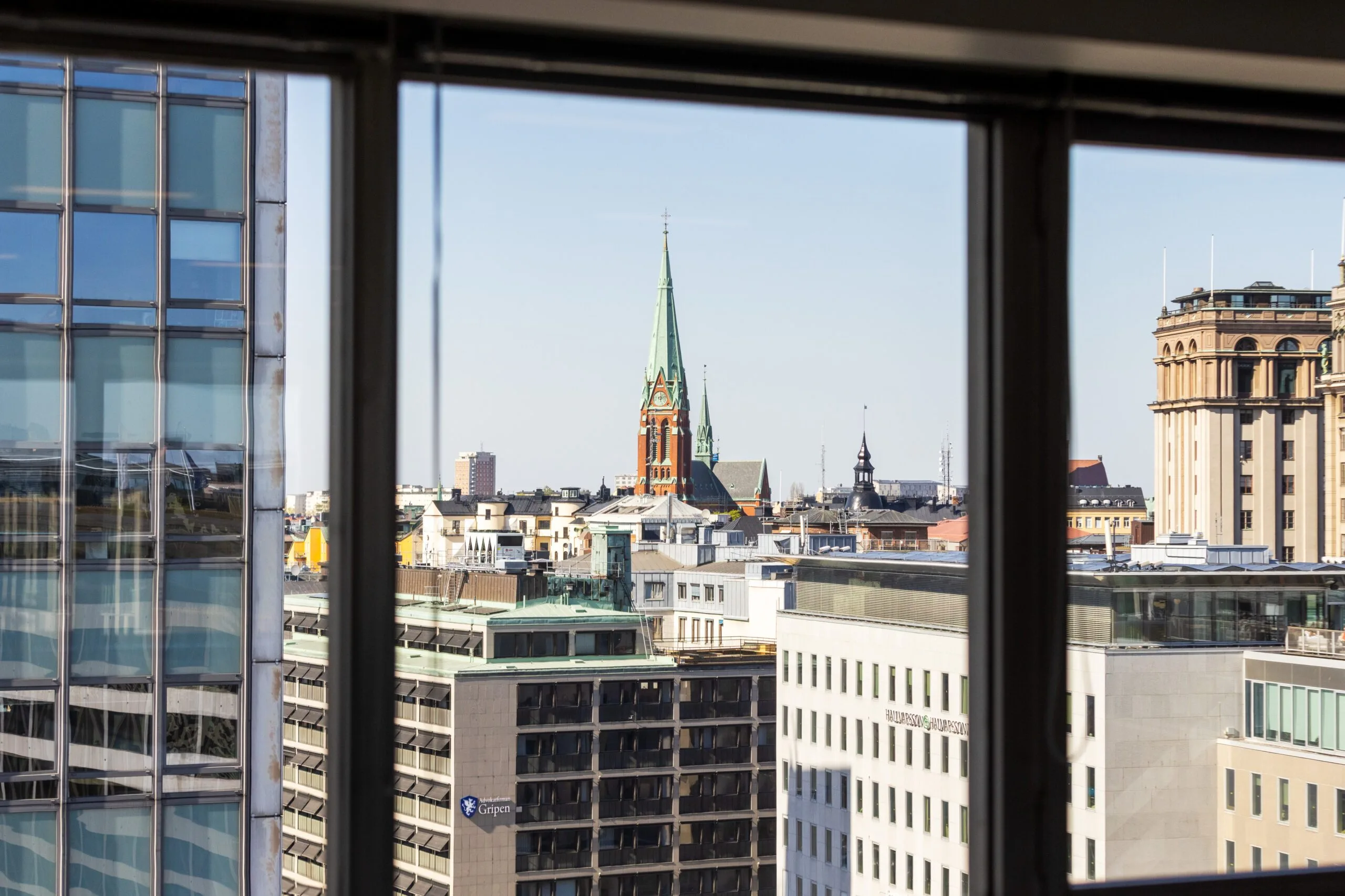 Utsikt genom ett fönster som visar en stadsmiljö med en kyrka med grön spira och omgivande byggnader.