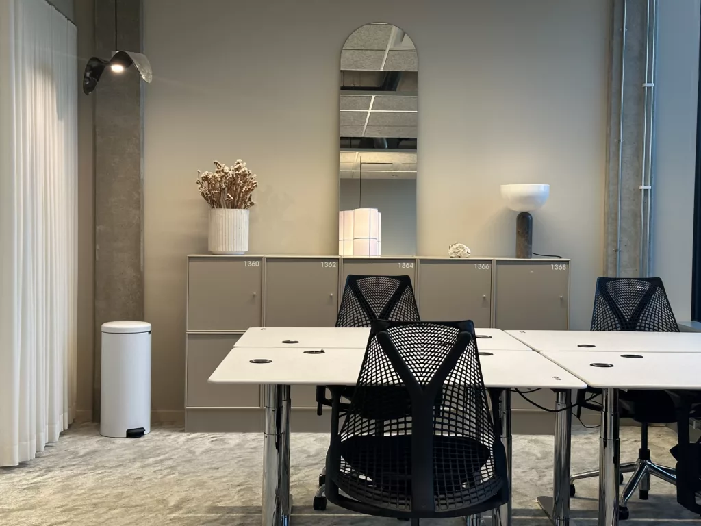 Ett kontorsutrymme med skrivbord, svarta stolar, en spegel och en hylla med en vas och lampa.