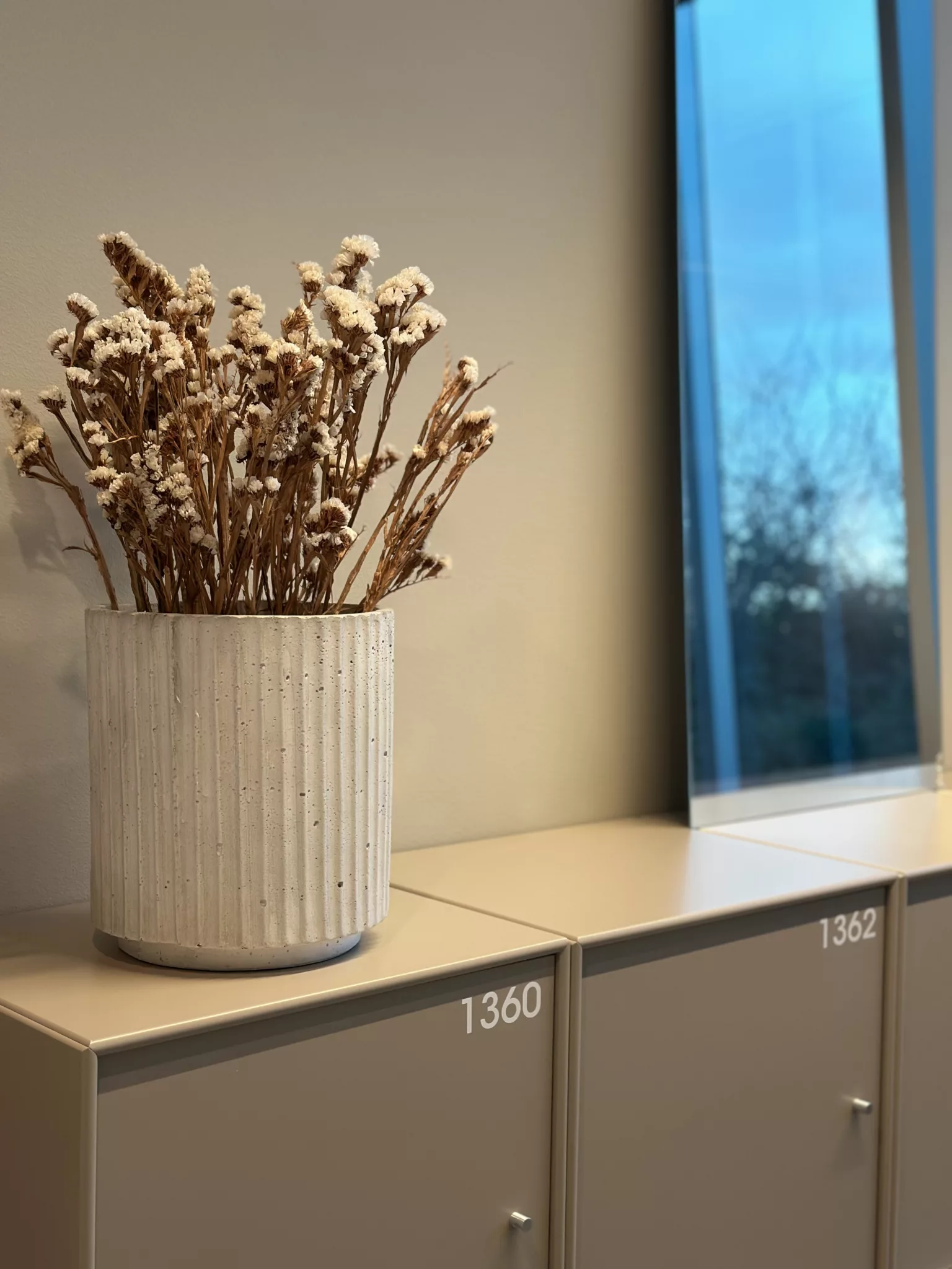 En vit vas med torkade blommor står på en ljus hylla bredvid ett fönster.