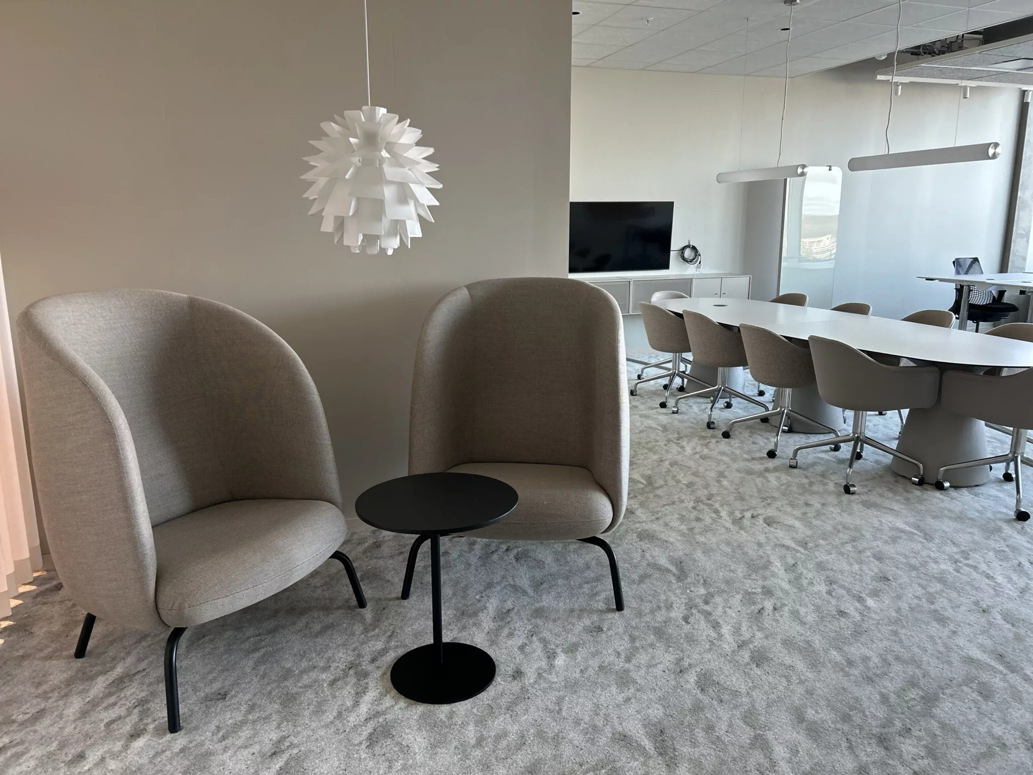 Två beige fåtöljer med ett svart litet bord mellan sig, en dekorativ vit taklampa, och ett konferensrum med bord och stolar i bakgrunden.