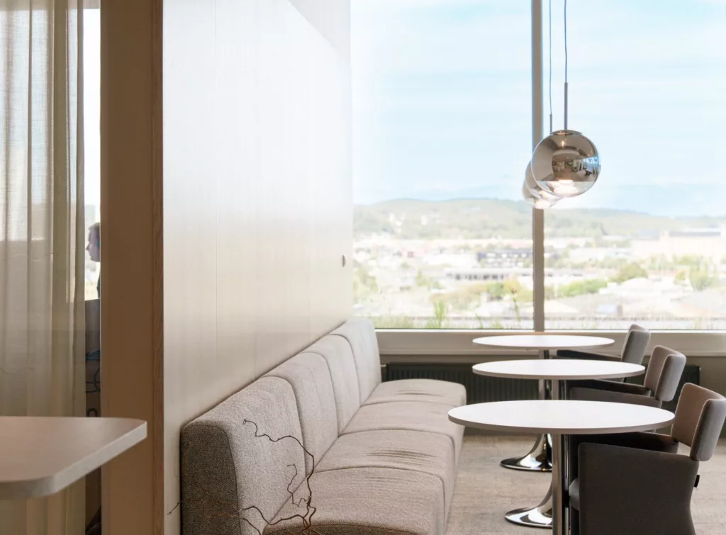 En lounge med beige soffa och små runda bord vid stora fönster som vetter mot en ljus utsikt.