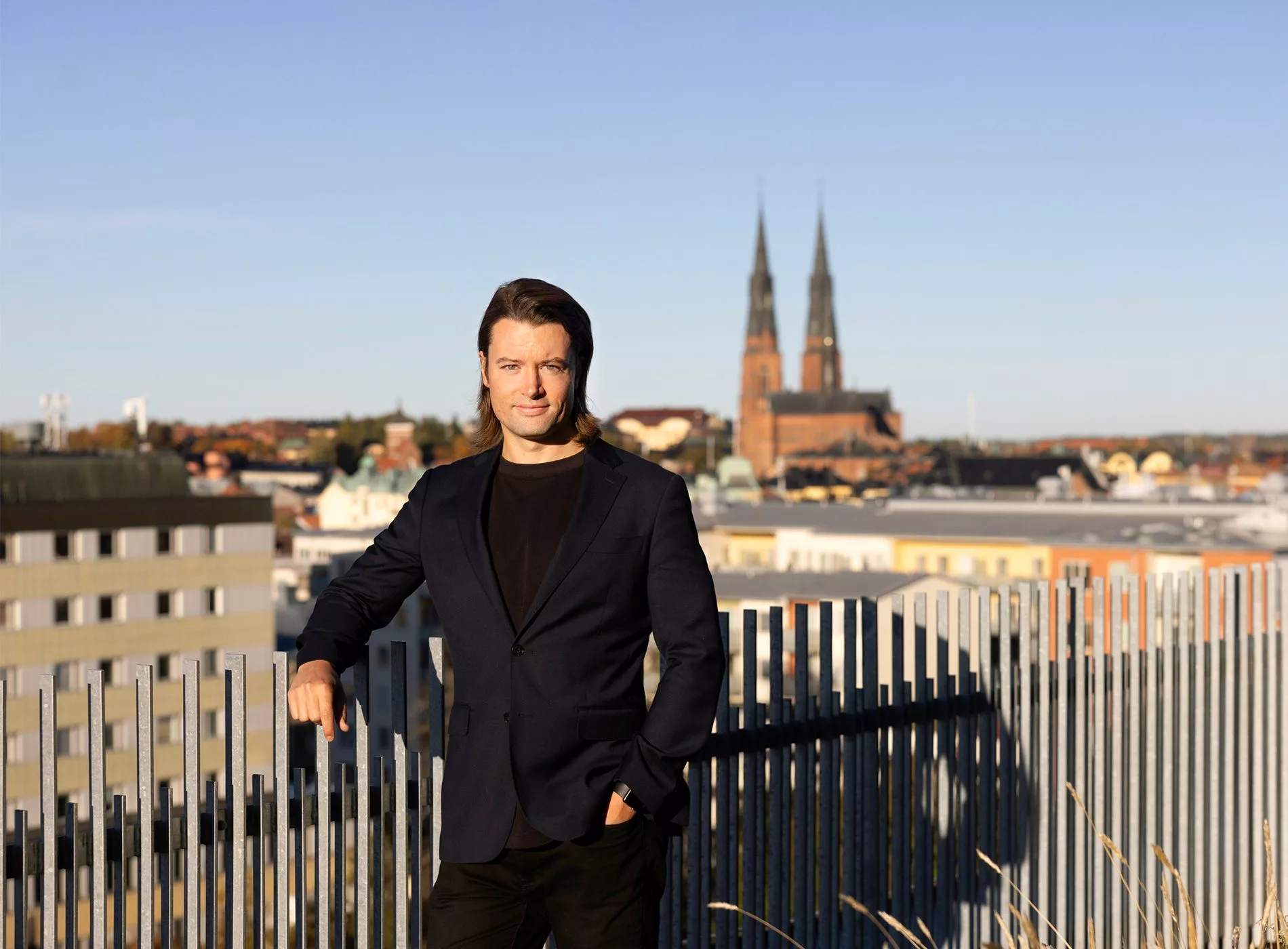 En man i mörk kavaj står framför ett staket med en stad och kyrkspiror i bakgrunden.