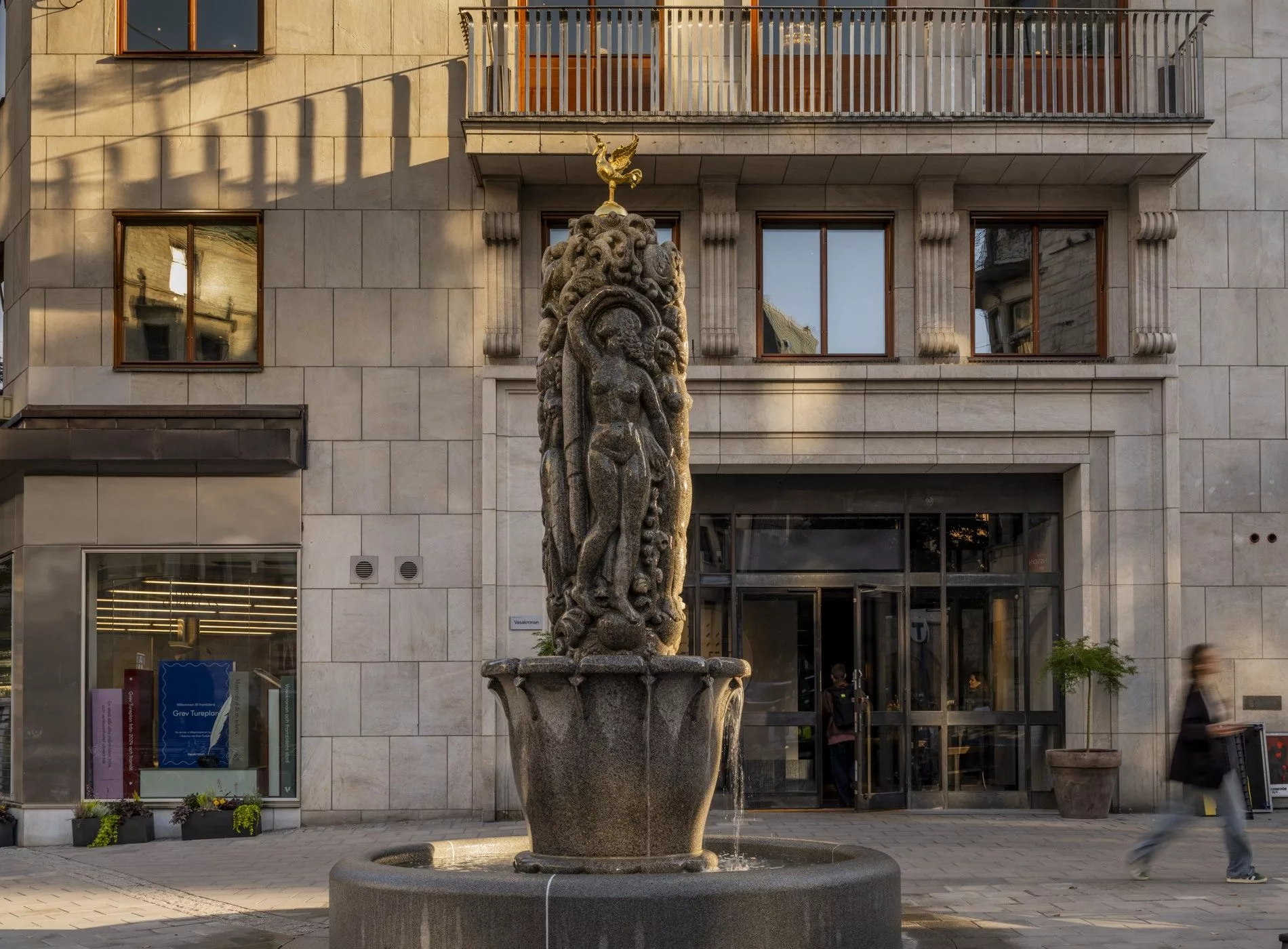 En detaljerad skulptur placerad i en fontän framför en byggnad med stora fönster.