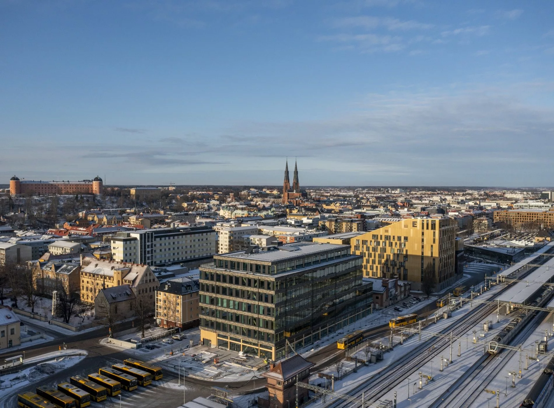 Uppsala stad i vintertid med snötäckta byggnader, kyrktorn i bakgrunden och järnvägsspår i förgrunden.
