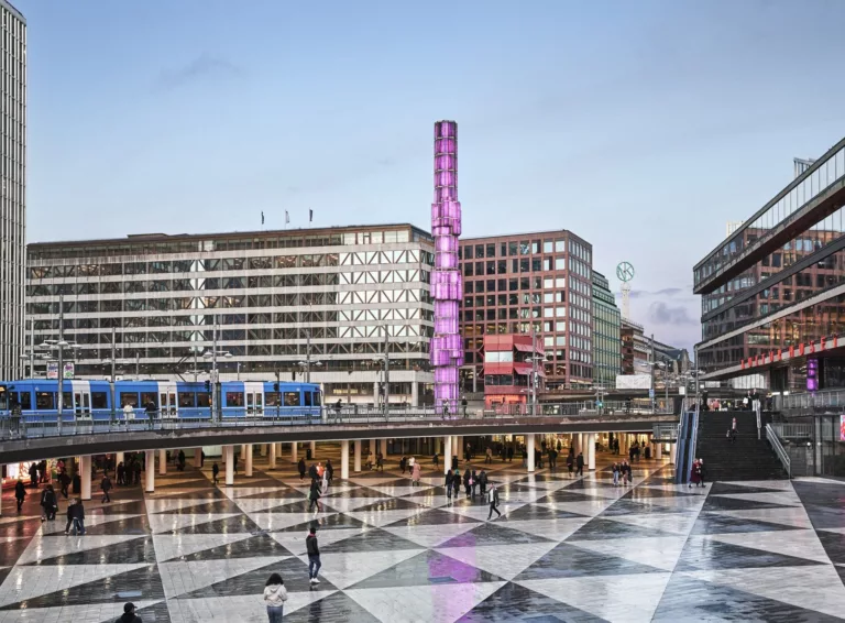 Sergels torg med svartvitt rutmönster på marken, en rosa glasobelisk och omgivande byggnader i kvällsljus.