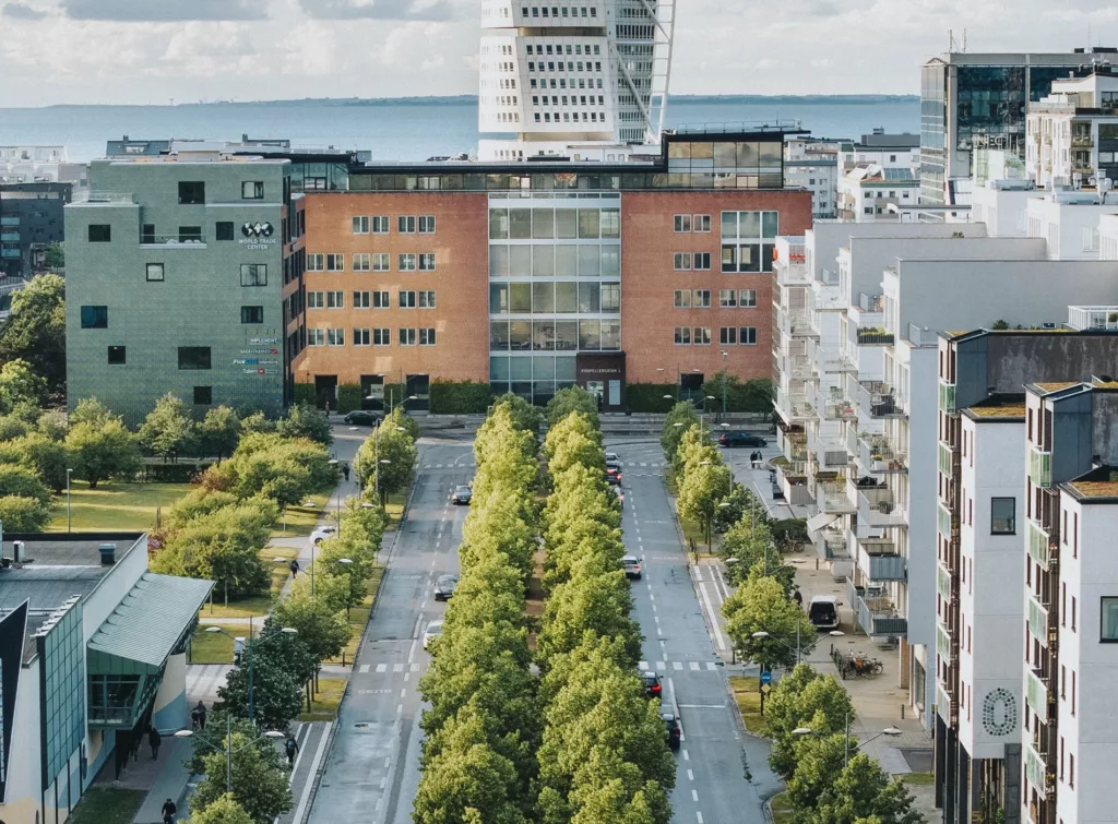 En grönskande allé med träd som leder till en röd byggnad, omgiven av moderna hus med Turning Torso i bakgrunden.