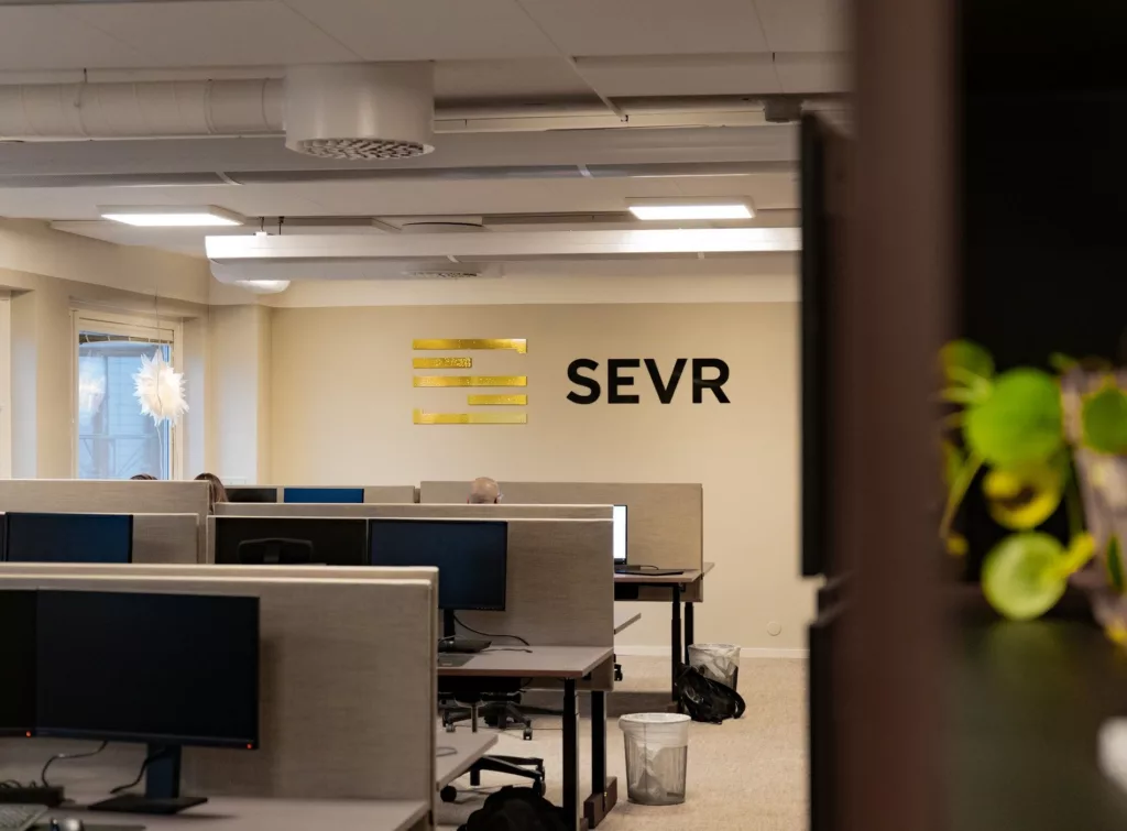 Ett kontorsutrymme med flera skrivbord och datorer, samt texten "SEVR" på väggen.