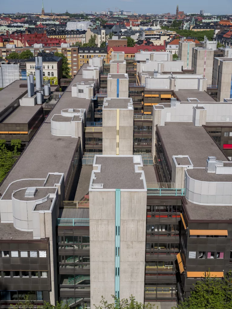 Flygfoto över en byggnad med modern arkitektur, omgiven av andra byggnader och grönska.