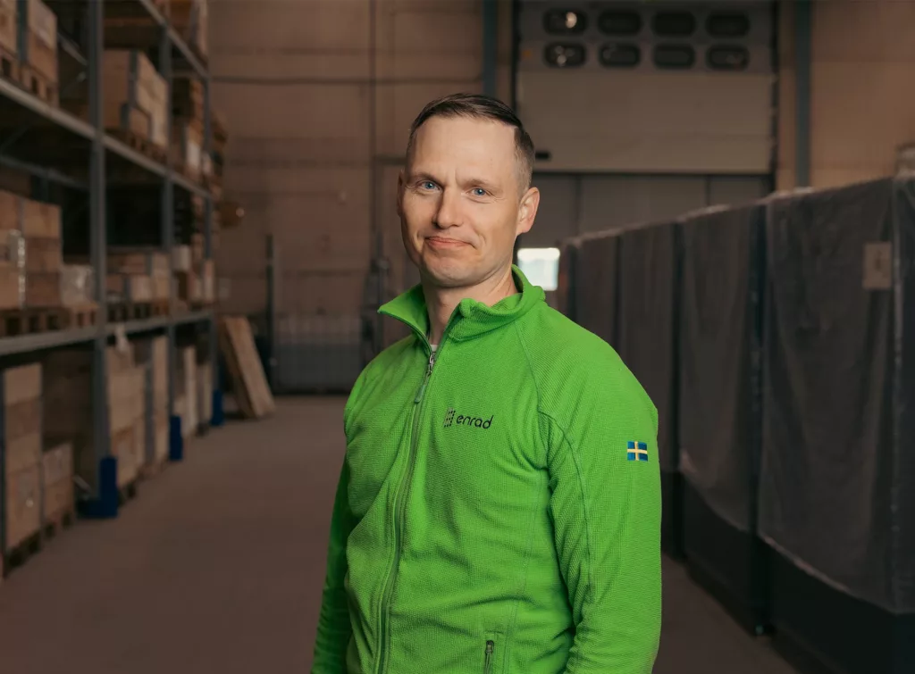 En man i grön jacka står inomhus bredvid lagerhyllor.
