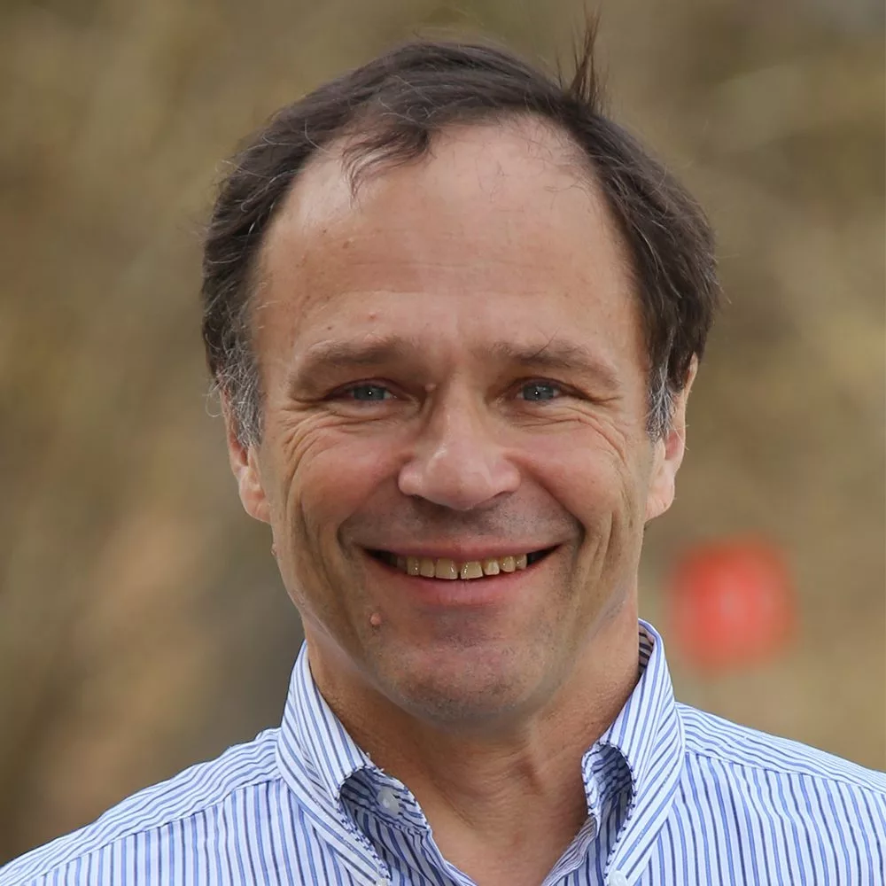 En man med blå- och vitrandig skjorta ler framför en oskarp bakgrund utomhus.