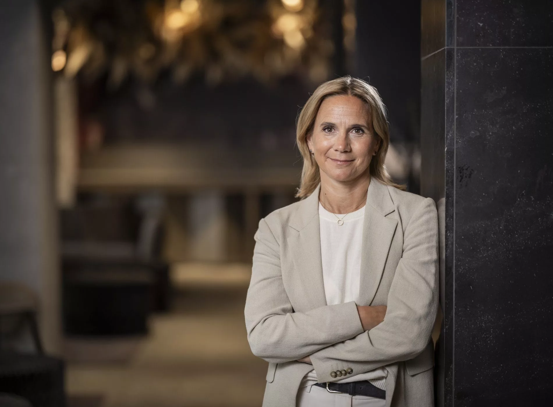 En kvinna i ljus kavaj står med armarna i kors i en elegant inomhusmiljö.