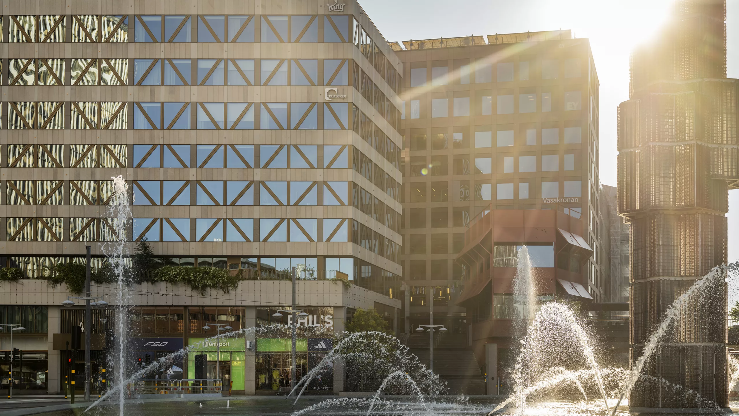En byggnad med glasfasad och fontäner framför, belyst av solljus.