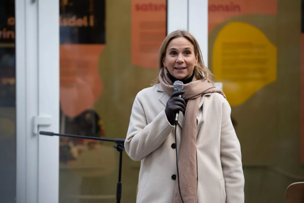 En person står utomhus och håller en mikrofon; bakgrunden visar skyltfönster med text.