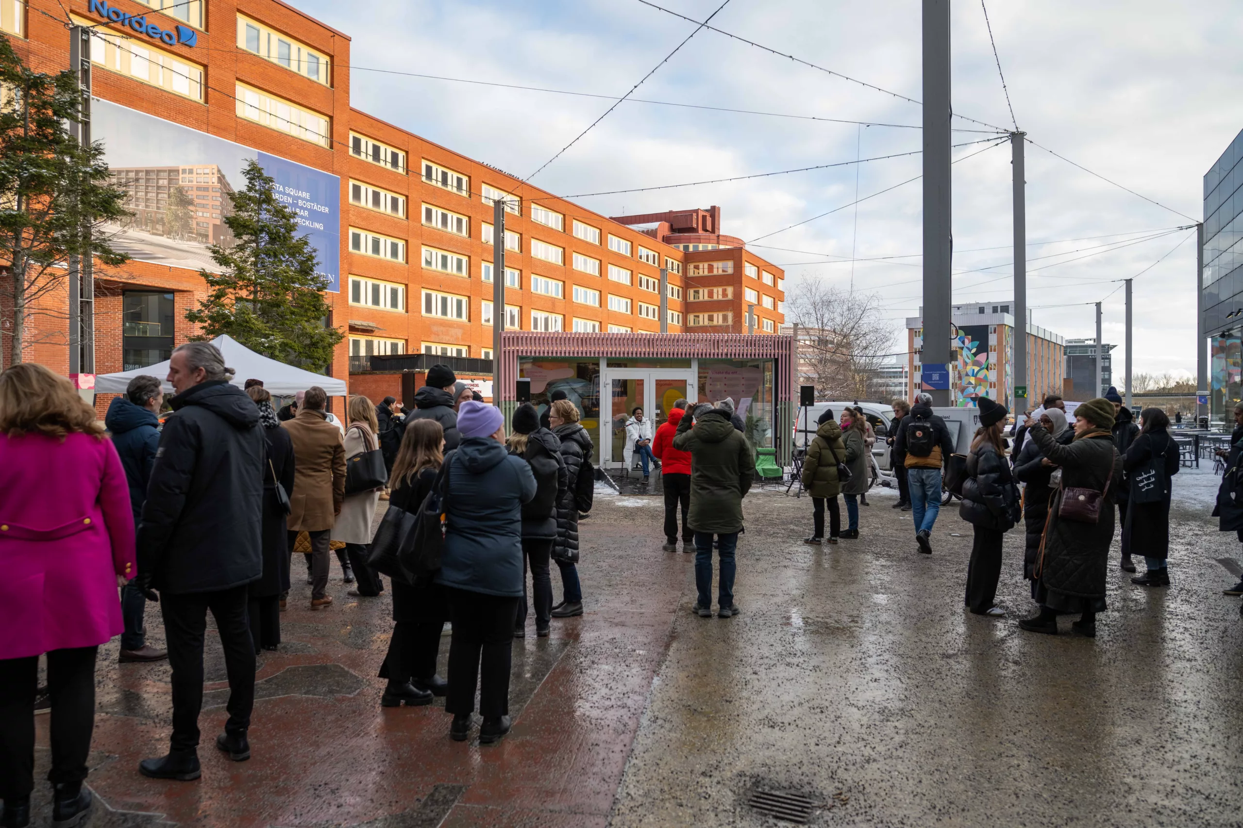 En grupp människor står utomhus på en trottoar nära byggnader med orange fasader under en molnig himmel.
