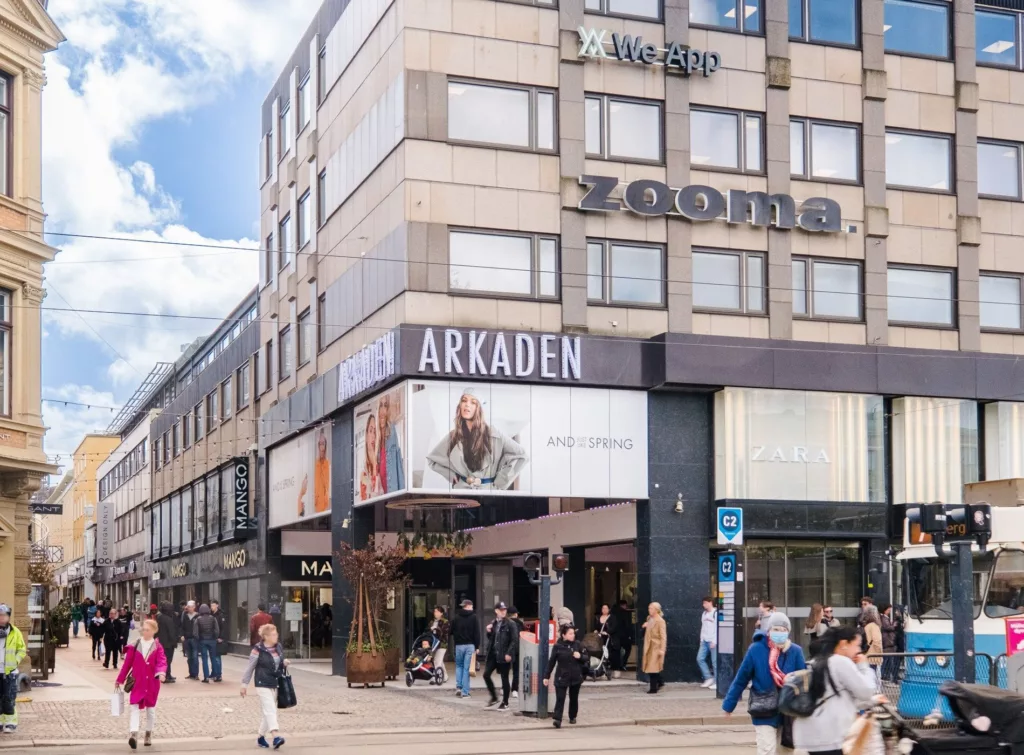 En byggnad med skylten "Arkaden" och "Rooms", omgiven av gående människor och butiker.