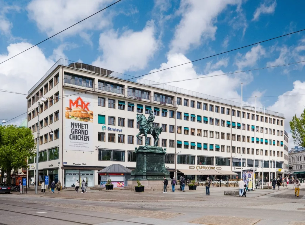 En stor byggnad med skylten "MAX Burgers", en staty av en ryttare och människor som promenerar på torget.
