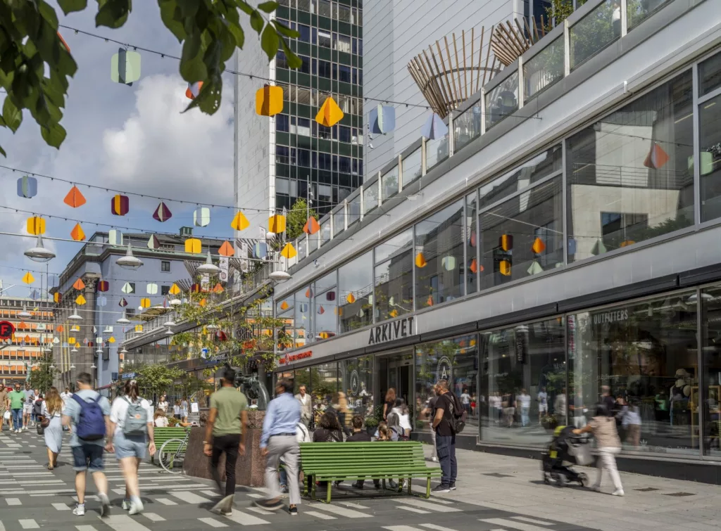 Fotografi av en stadsgata med människor, dekorationer och bord utomhus, omgiven av moderna byggnader.