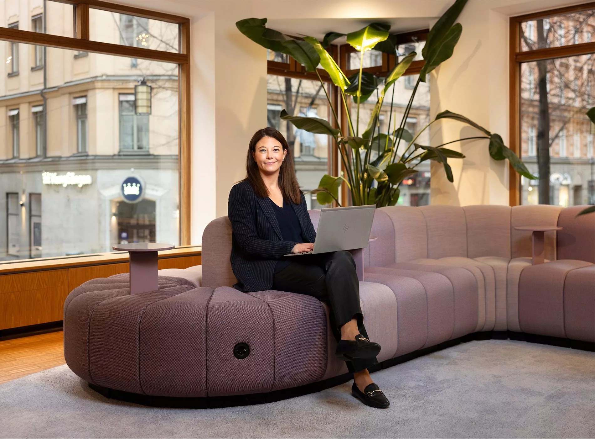 En kvinna sitter på en lila soffa med en bärbar dator i ett kontor med stora fönster och gröna växter.