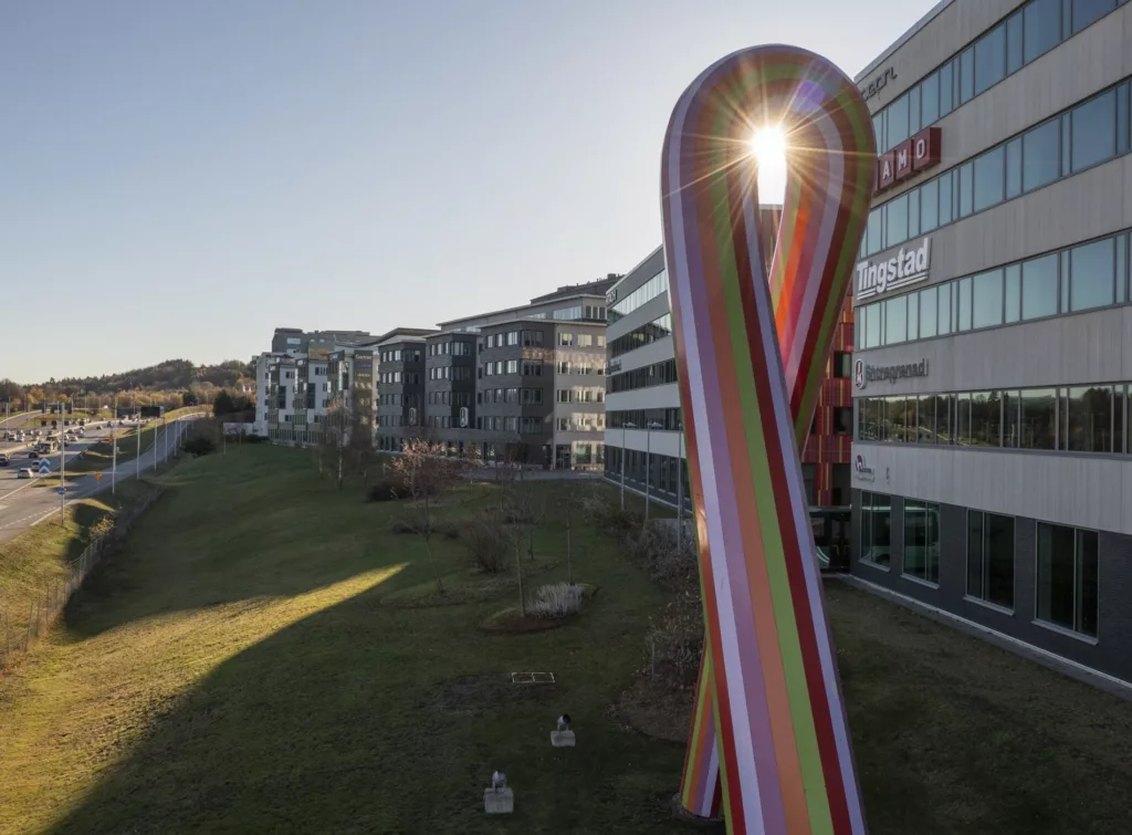En stor färgglad skulptur i form av ett band står framför en rad moderna byggnader, med solens ljus som skiner igenom den.