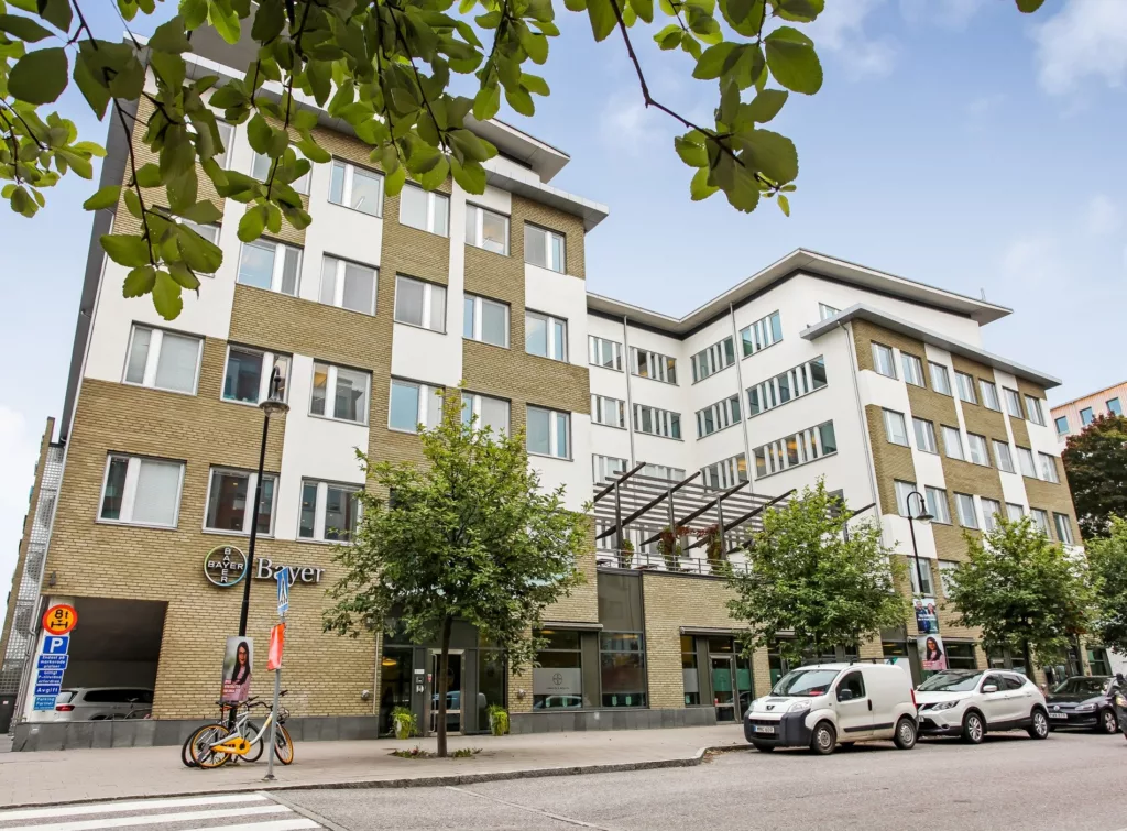 En kontorsbyggnad med vit och beige fasad, omgiven av träd, cyklar och parkerade bilar.