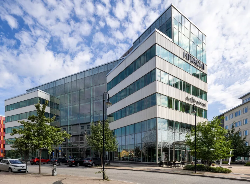 En modern glasbyggnad med flera våningar under en blå himmel med moln, omgiven av träd och trottoarer.