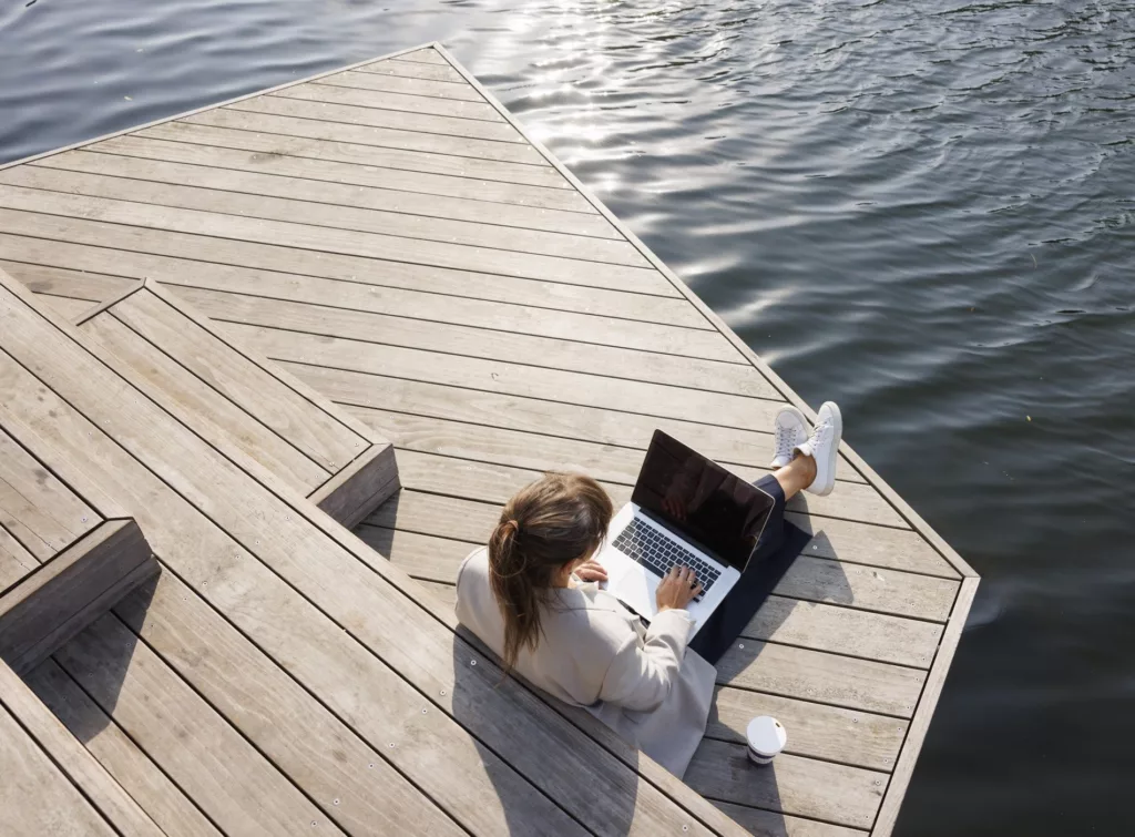 En person sitter på en brygga vid vatten och arbetar på en laptop.