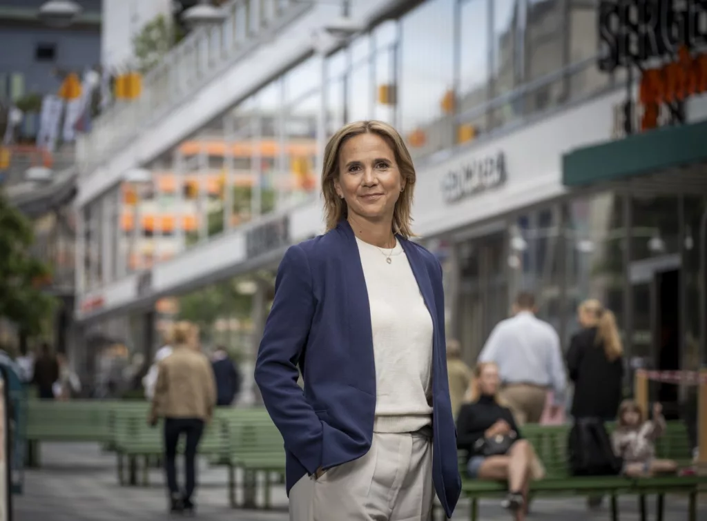 En kvinna i blå kavaj står utomhus på en livlig gata med butiker och människor i bakgrunden.