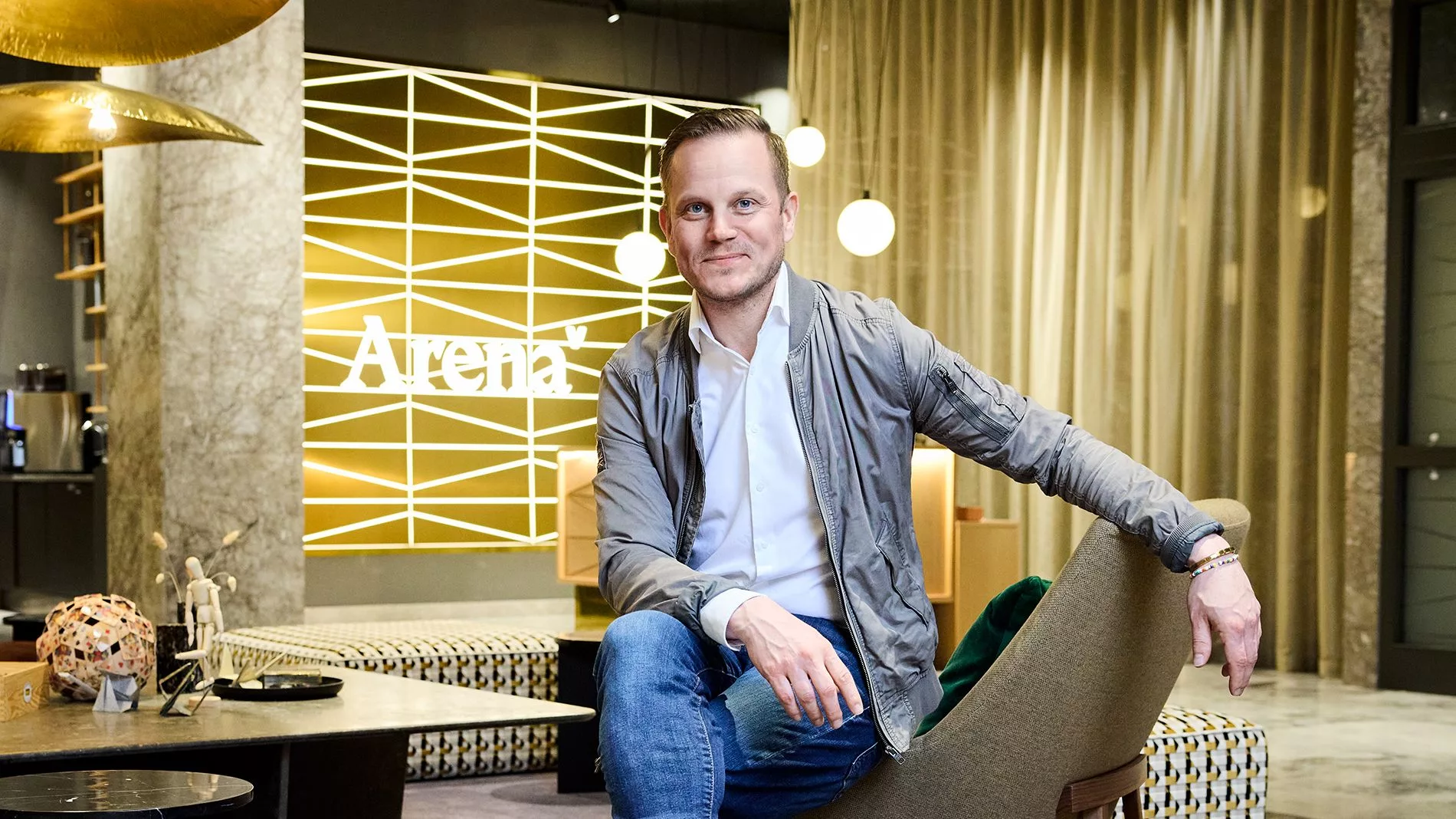 En man i grå jacka och blå jeans sitter avslappnat i en modern miljö med texten "Arena" på väggen bakom honom.