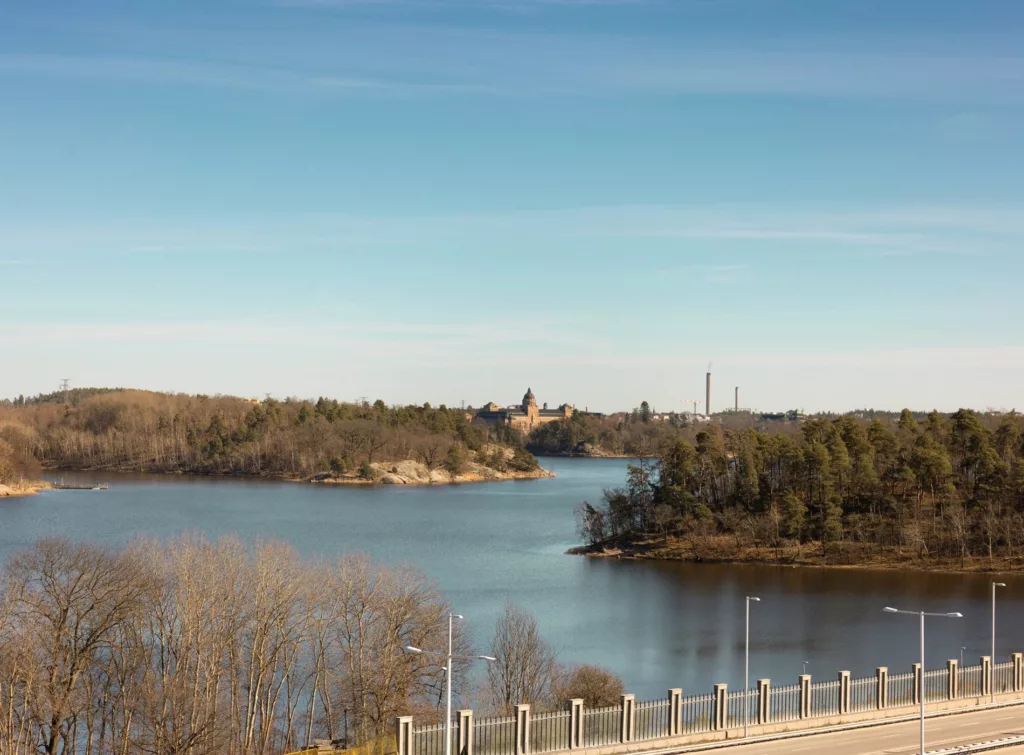En sjö med omgivande skog och byggnader i bakgrunden under klarblå himmel.