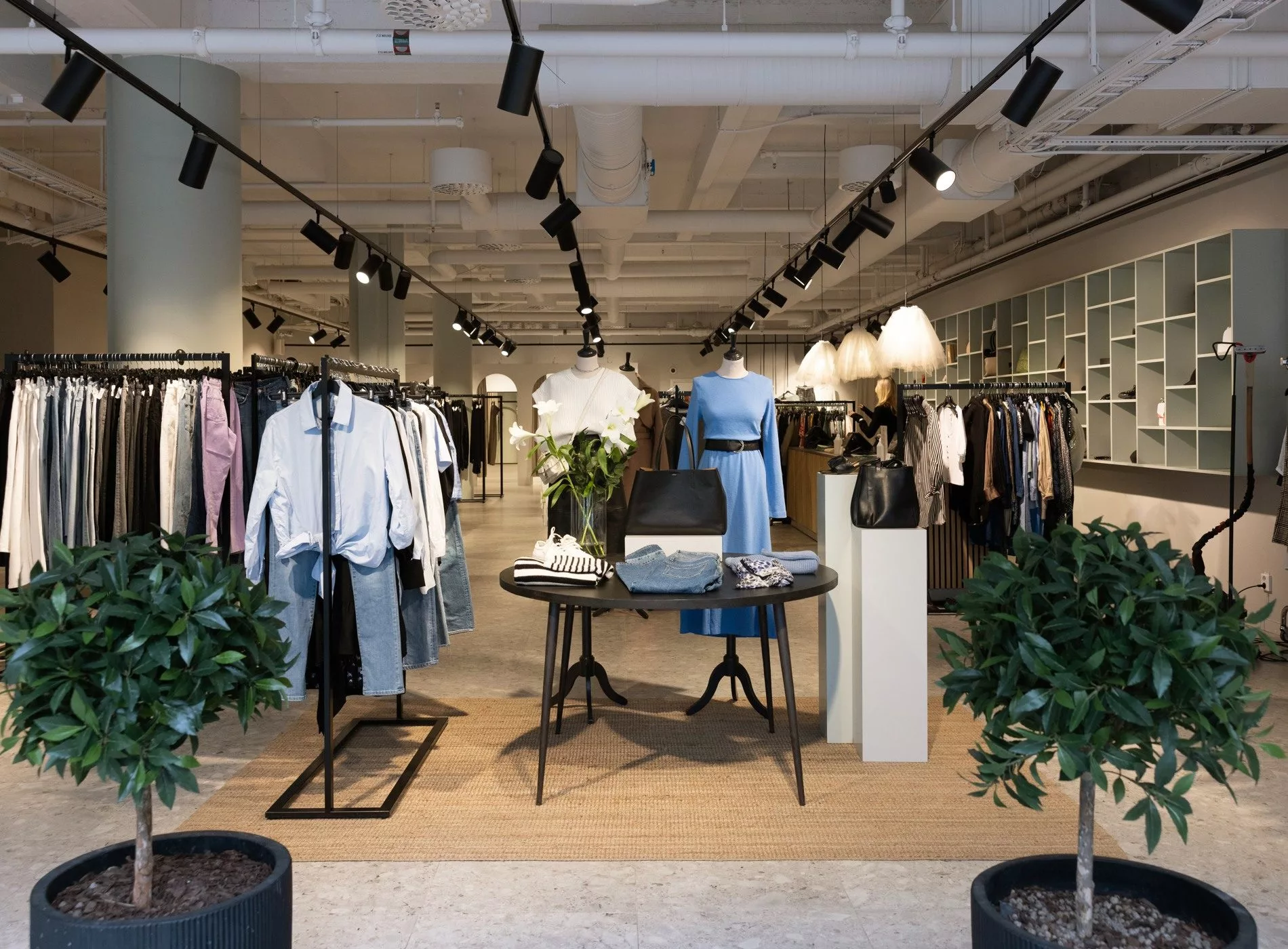 En butik med klädställningar fyllda med plagg, bord i mitten och dekorativa växter.