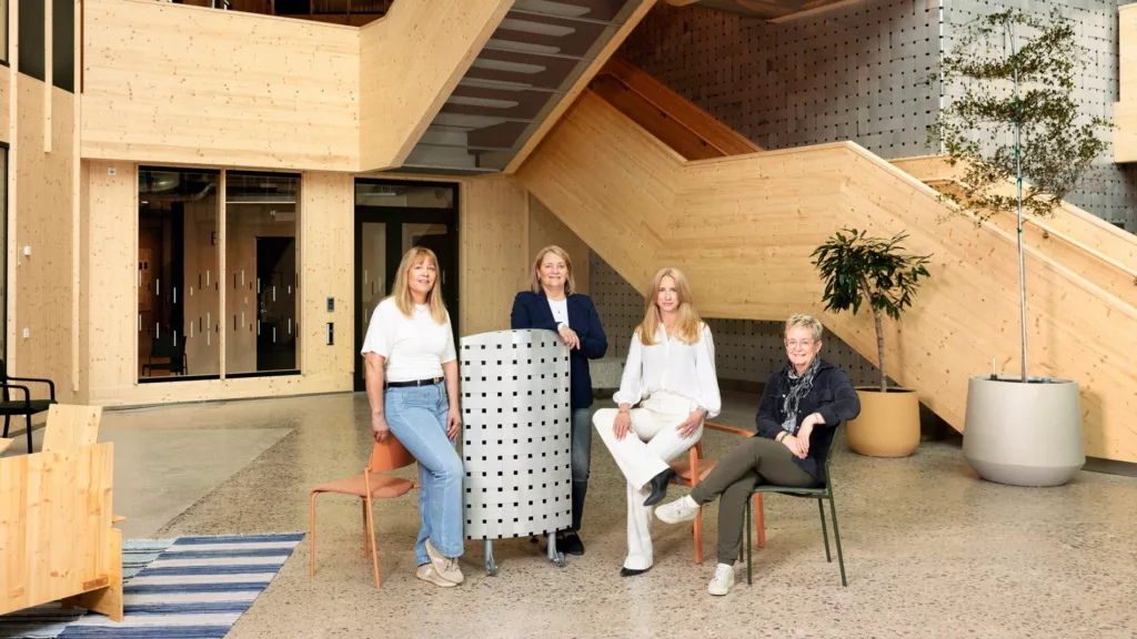 Fyra personer står och sitter runt en möbel i en ljus entré med trappor och växter i bakgrunden.
