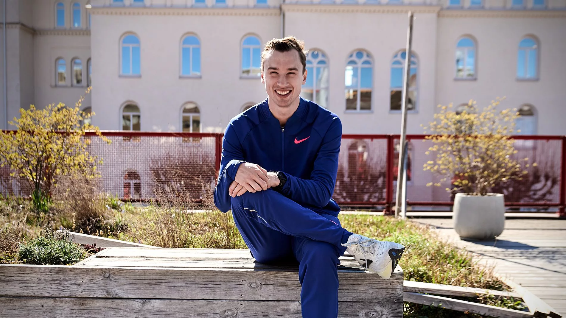 En ung man i blå träningskläder sitter utomhus på en träbänk, med en byggnad i bakgrunden.