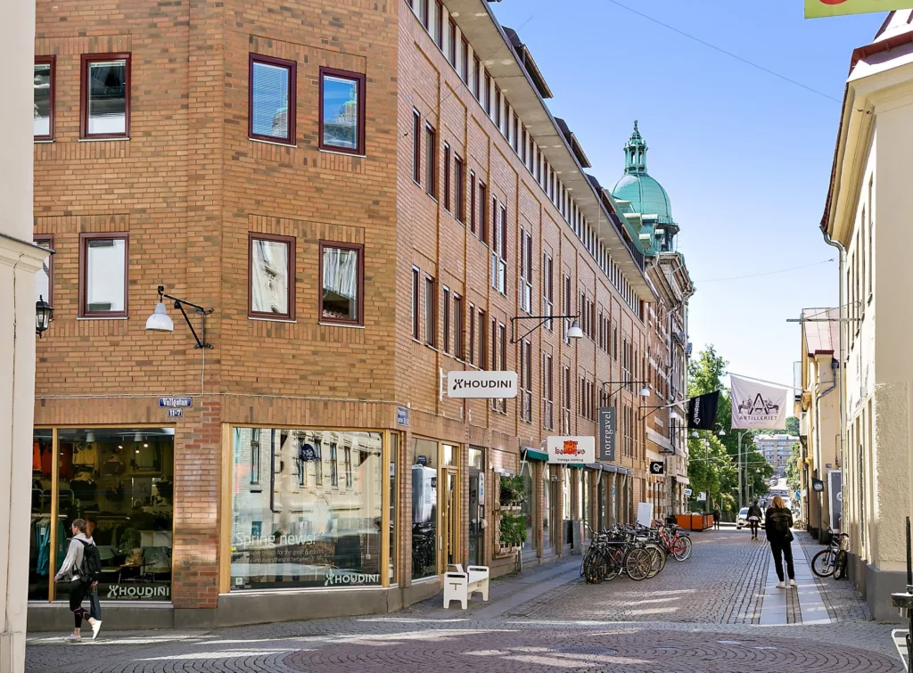 En gata med tegelbyggnader, butiker och cyklar, med en grön byggnadsdetalj synlig längre bort.