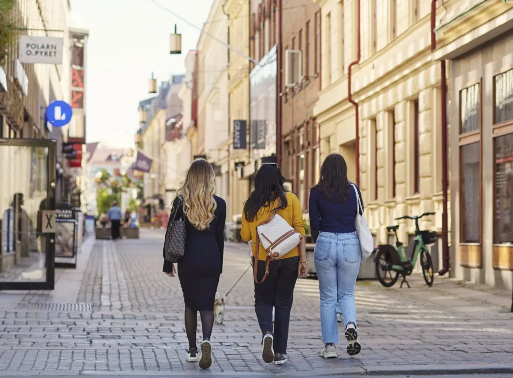 4. Tre kvinnor går på en solig stadsgata med cyklar parkerade vid sidan och äldre byggnader i bakgrunden.