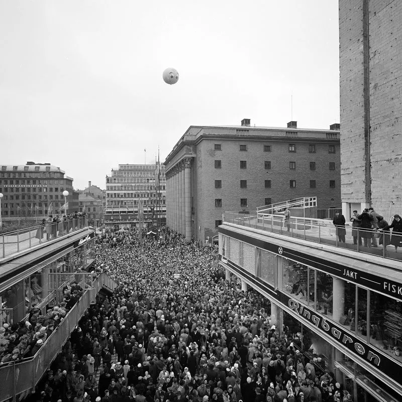 Svartvit bild av en stor folkmassa på ett torg, omgiven av byggnader och gångbroar.