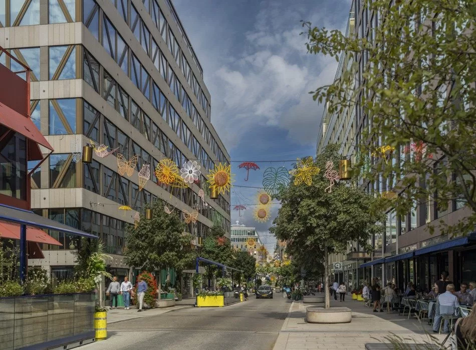 En stadsgata kantad av moderna kontorsbyggnader, träd och färgglada dekorationer hängande ovanför vägen under en blå himmel.