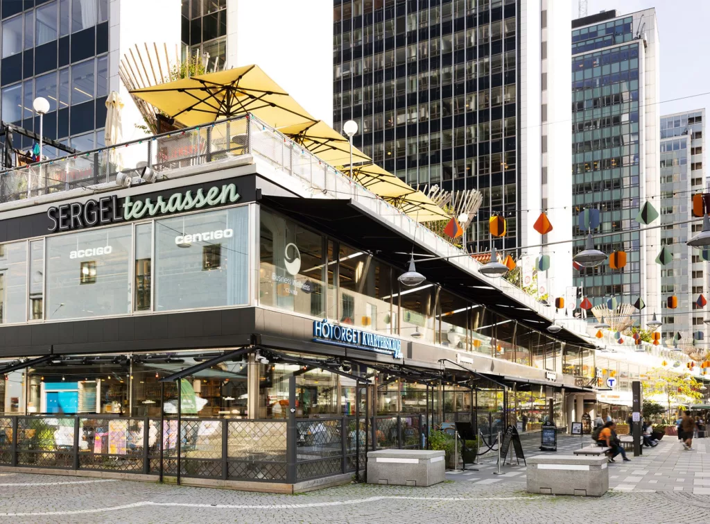 Restaurang med skylt "Terrassen" och uteservering under gula parasoller, omgiven av moderna byggnader.