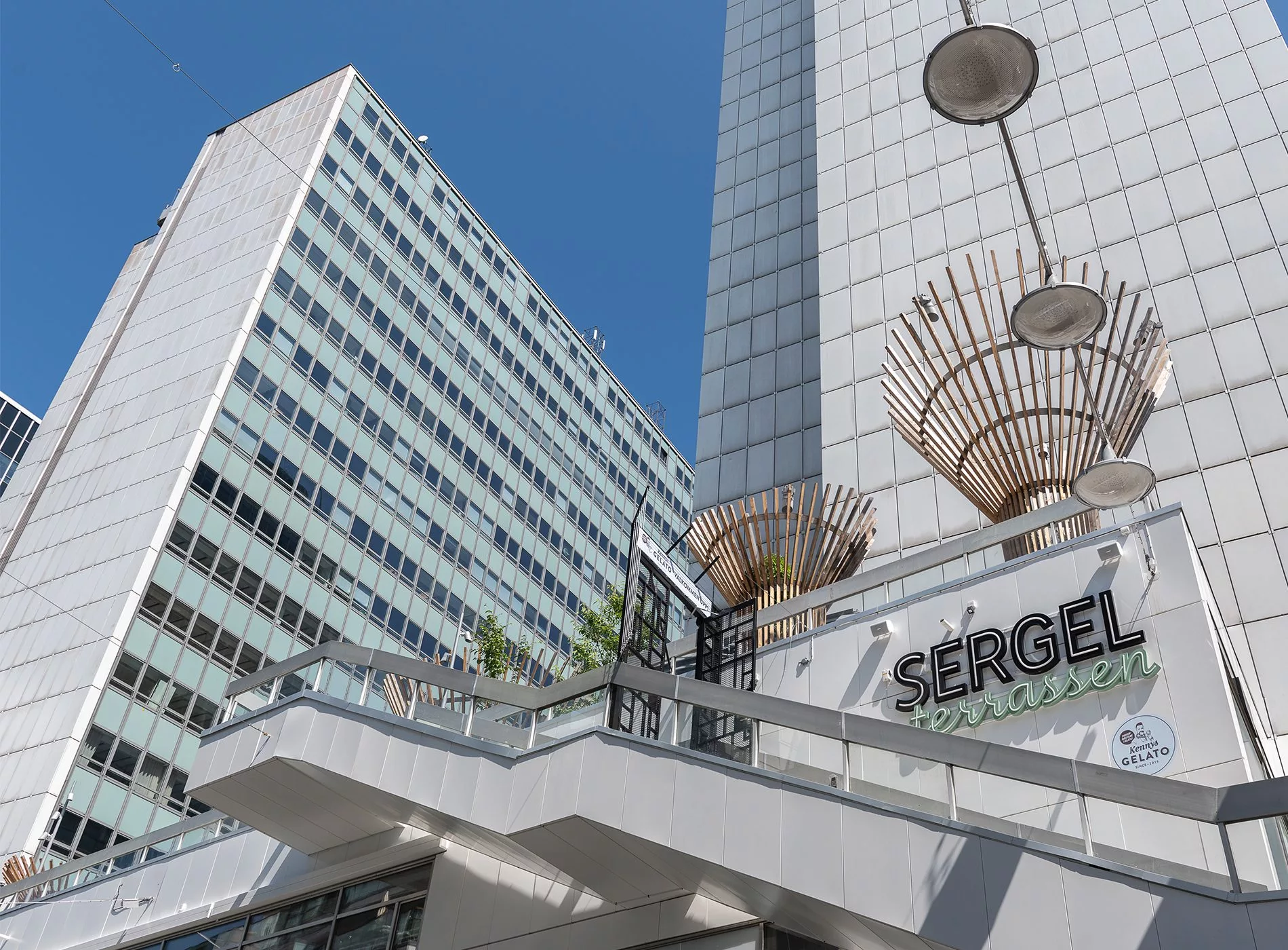 Byggnader med texten "Sergel" och stora dekorativa ljusarmaturer mot en blå himmel.
