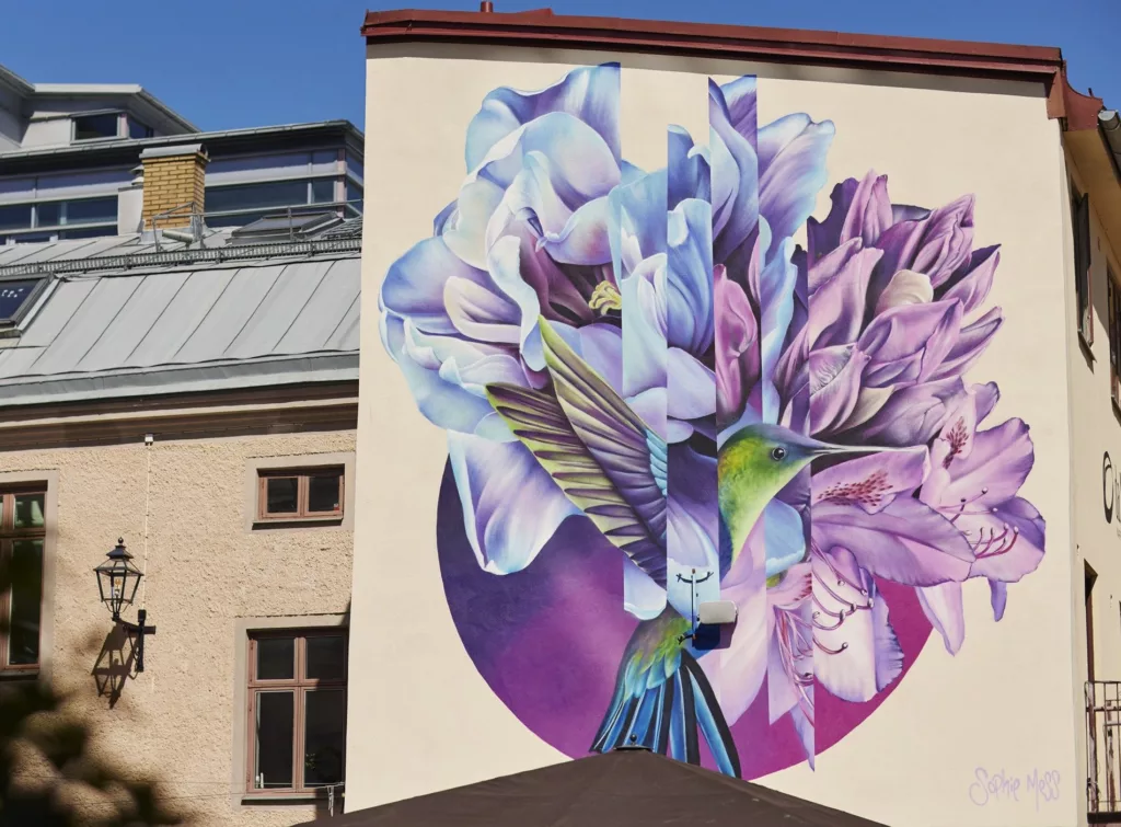 En väggmålning av en färgglad blomma och en kolibri på en byggnad.