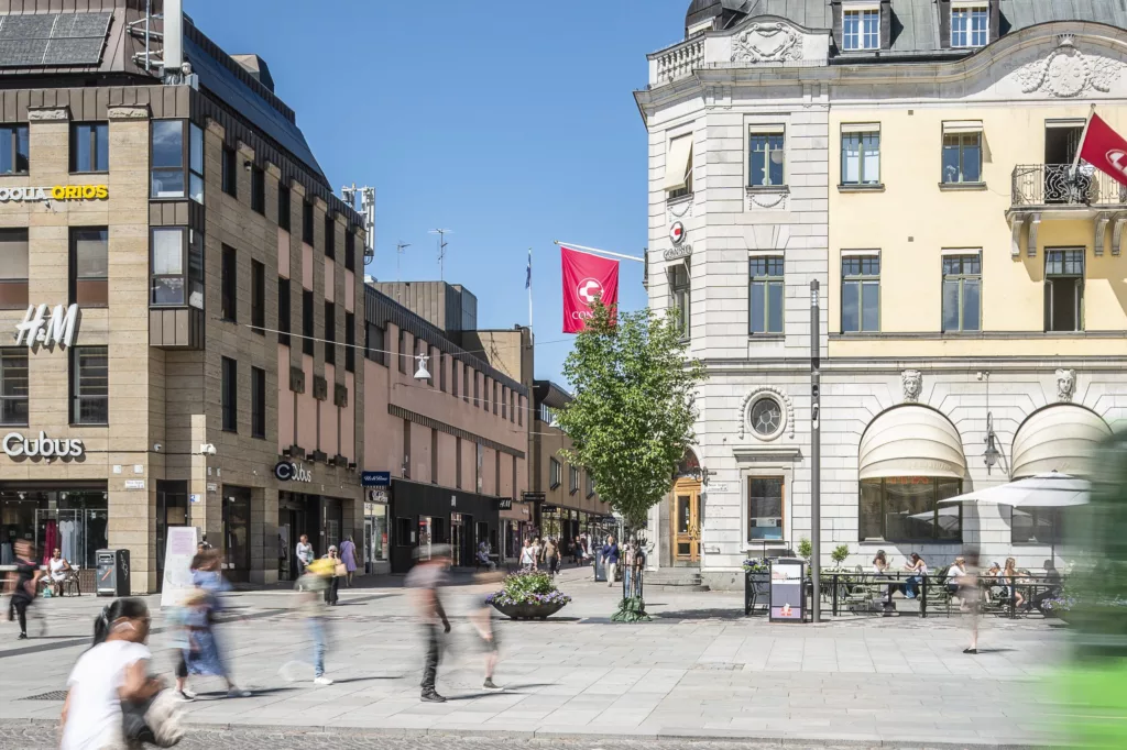 Ett öppet torg med människor som promenerar, omgivna av äldre byggnader och en röd flagga på en flaggstång.